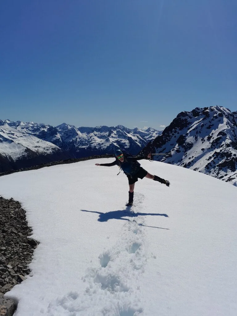 A hiker in alpine gear stands on snow covered ground with an ice axe with the Spencer Range in the background on the climb to Princess Bath in St James Conservation Area