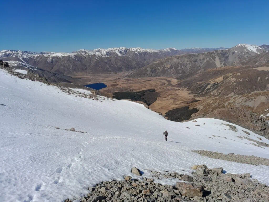 A hiker ascends a steep snowy slope with Lake Tennyson in the background a snowy covered Crimea Range on the skyline