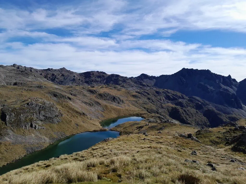 A view of a blue tarn, Hinapouri tarn, amongst tussock, on the climb to Angelus Peak, with Bristol pass in the background
