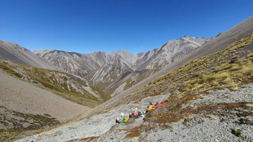 A group of trampers sitting on a high mountain ridge for lunch at Inkerman Saddle, overlooking the glaciated valley and scree peaks of the Two Thumb Range in Canterbury. A view towards Brabazon Saddle in the distance