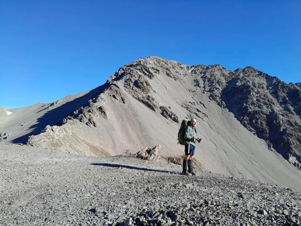 A tramper stands on a high alpine saddle with rugged peak called Graf Spee behind him in the Two Thumb Range 