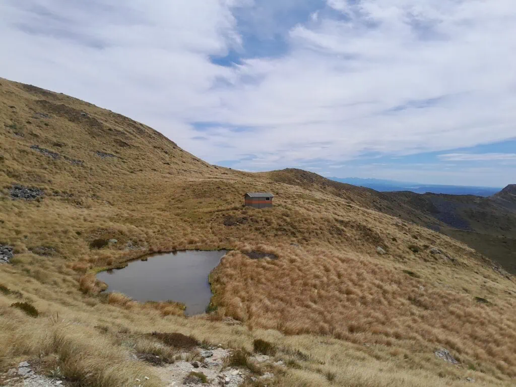 A small and cozy Mt Misery Hut amongst tussock next to a tarn with fantastic views across to the Travers Range