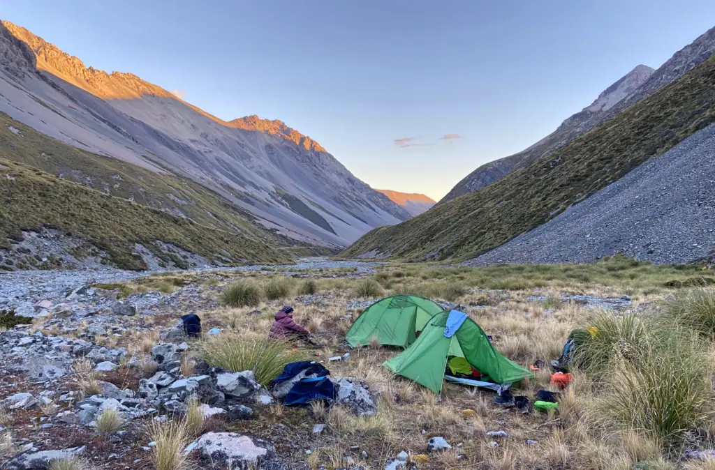 Backcountry camping in North East Gorge Stream, Two Thumb Range. Two green tents with one tramper are pitched in a tussock clearing as sunset light hits the surrounding scree mountain peaks in Canterbury, New Zealand. Stag saddle ridge of the Te Araroa Trail is visible in the distance