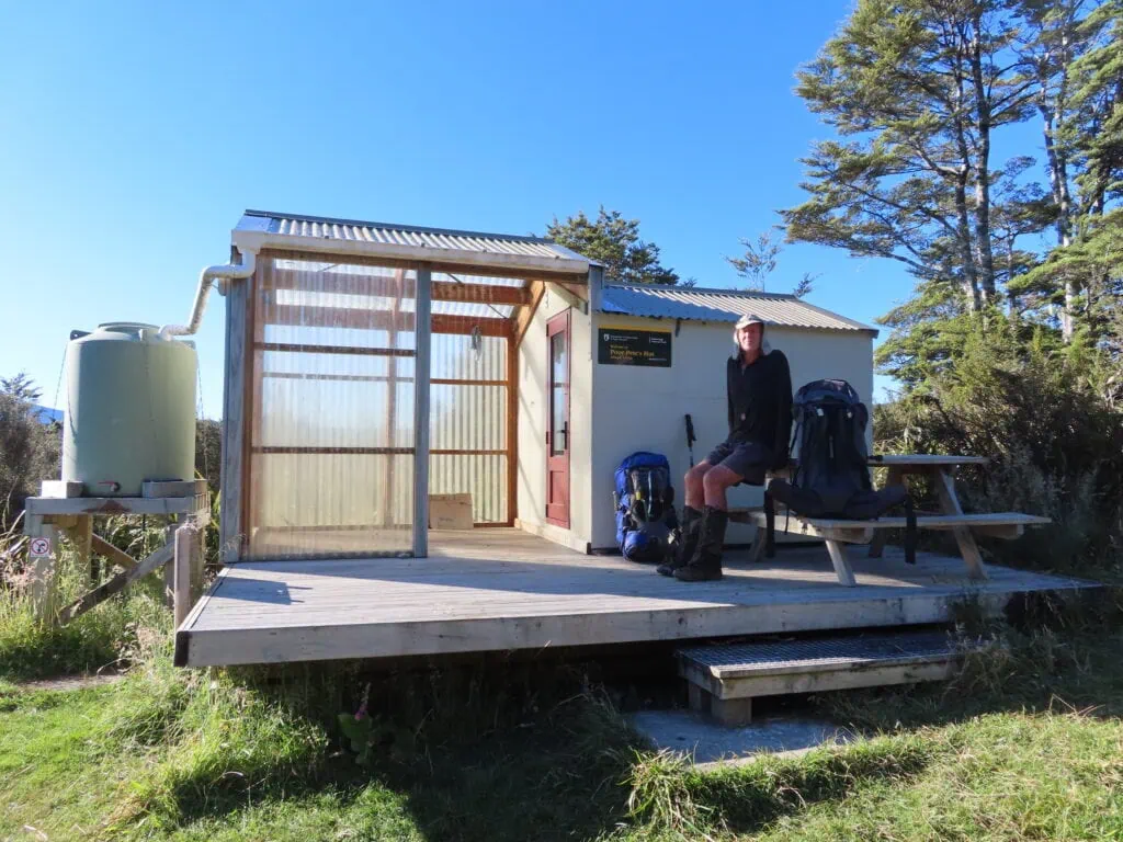 A hiker sits outside a small 2 person department of conservation hut called Poor Pete's Hut, under a blue sky on the 1000 Acre Plateau