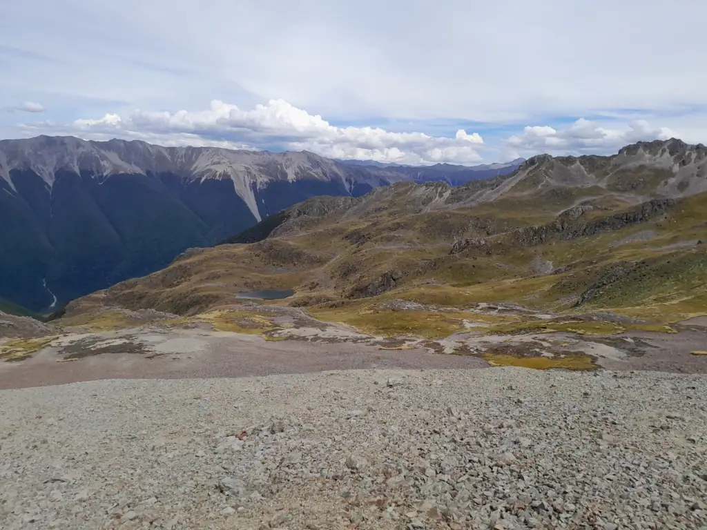 A view from Robert Ridge showing a scree slope leading down to a tarn nestled amongst alpine grass with St Arnaud Range in the distance