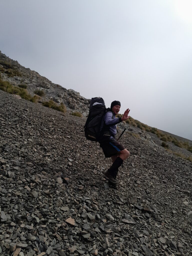 A tramper in a blue jacket descending a steep grey scree slope from Brabazon Saddle in the Two Thumb Range, Canterbury.