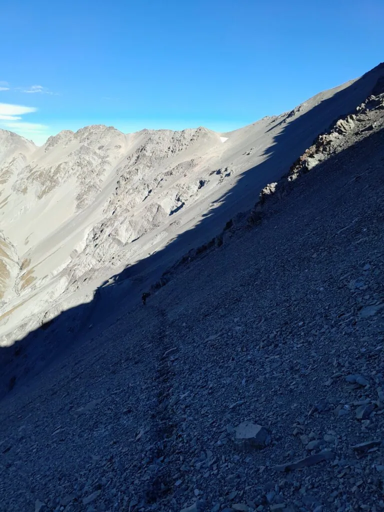A tramper ascending a steep, expansive scree slope to Balaclava Saddle in the Two Thumb Range. Plata Col is visible in the background. The landscape showcases the rugged alpine terrain of the Te Kahui Kaupeka Conservation Area, Canterbury, under a hazy sky.