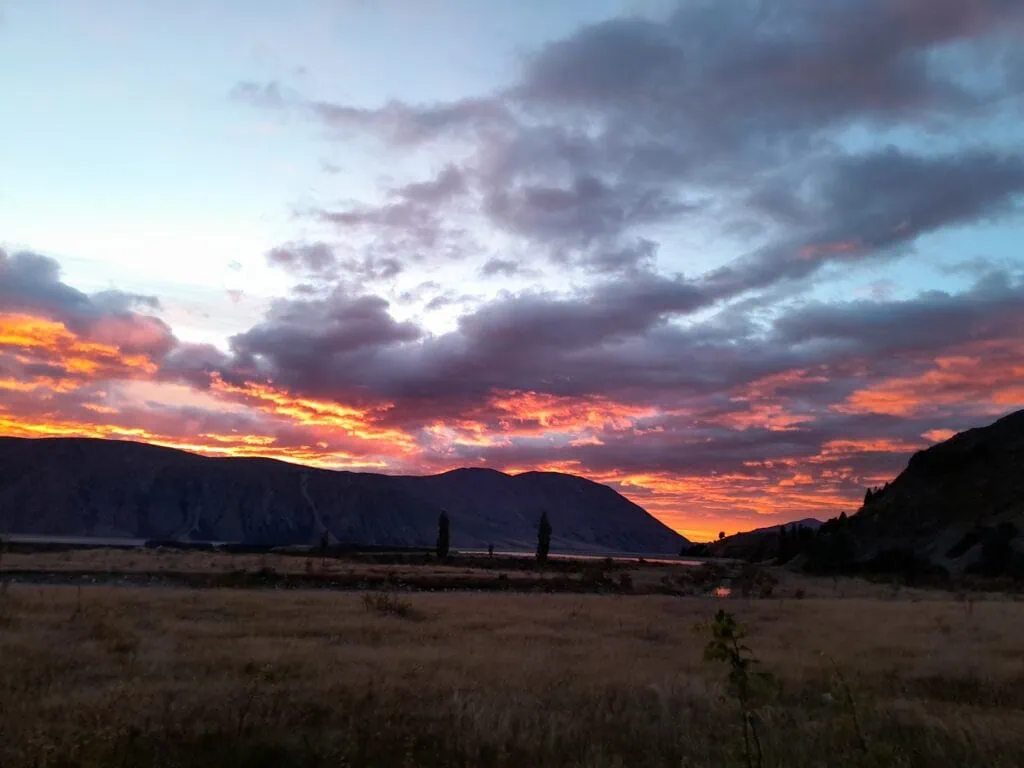 A vibrant orange and red sunrise over the Harper mountain ranges at the start of the Bush Stream at Mesopotamia in the Two Thumb Range. The landscape features silhouetted hills and golden tussock flats under a dramatic sky in the Canterbury high country