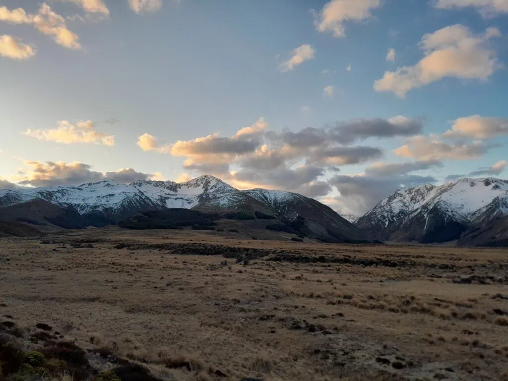 Tussock covered flats from Maling Pass Road in front of a snowy covered Mt Princess with clounds and the start of sunset in St James Conservation Area