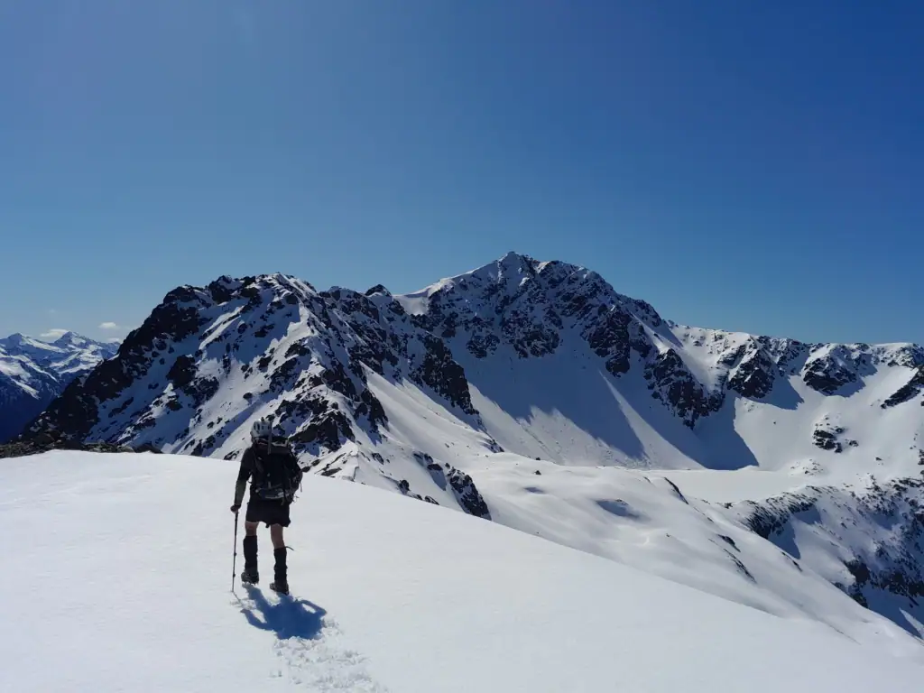 A hiker with alpine gear stands on a snowy point with a frozen snow-covered Princess Bath opposite him and Mt Princess in front, St James Conservation Area