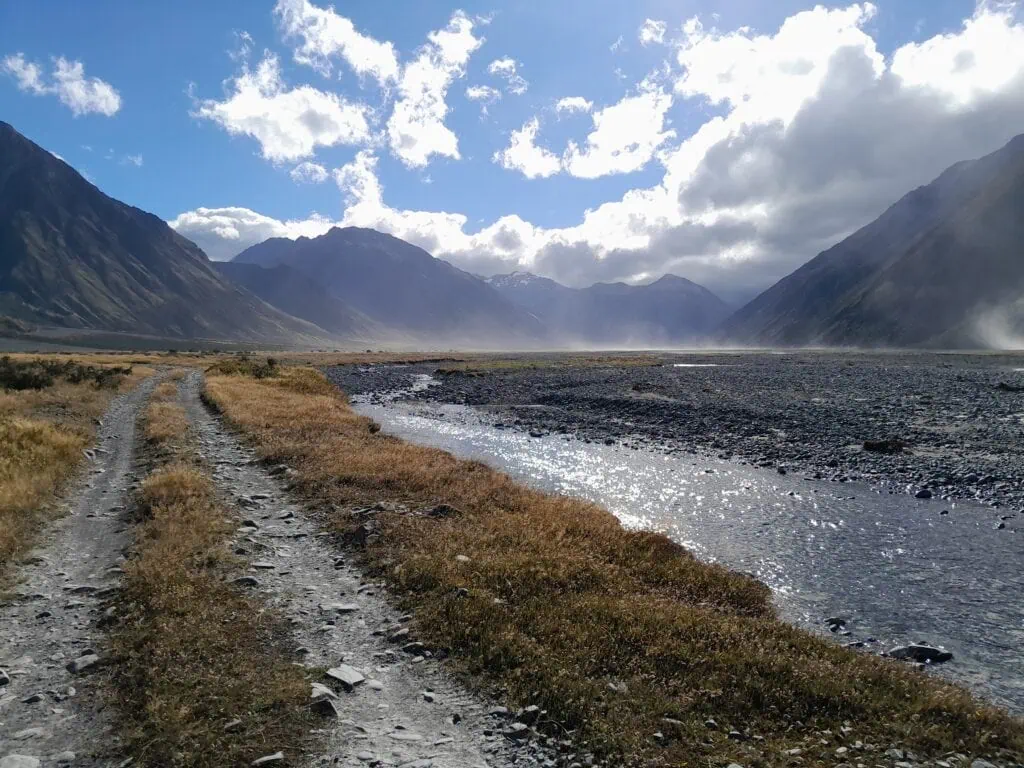 A 4WD track alongside the Havelock River at Mesopotamia, Canterbury. The image captures strong winds creating dust storms across the wide, braided riverbed, with the rugged mountains of the Two Thumb Range and Southern Alps in the background.