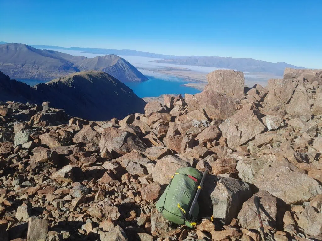 A small tramping pack with alpine gear including ice-axe attached sitting amongst boulders looking down to Lake Lake Ōhau on the way to Dumb-bell Lake