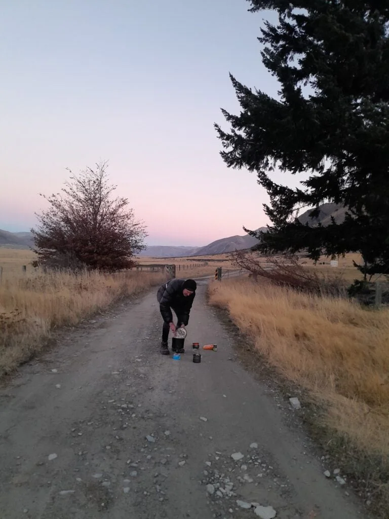 A hiker cooks dinner on a gas stove in the middle of a gravel road in Ahuriri Conservation Park near Birchwood Homestead at the base of Mt Gladwish