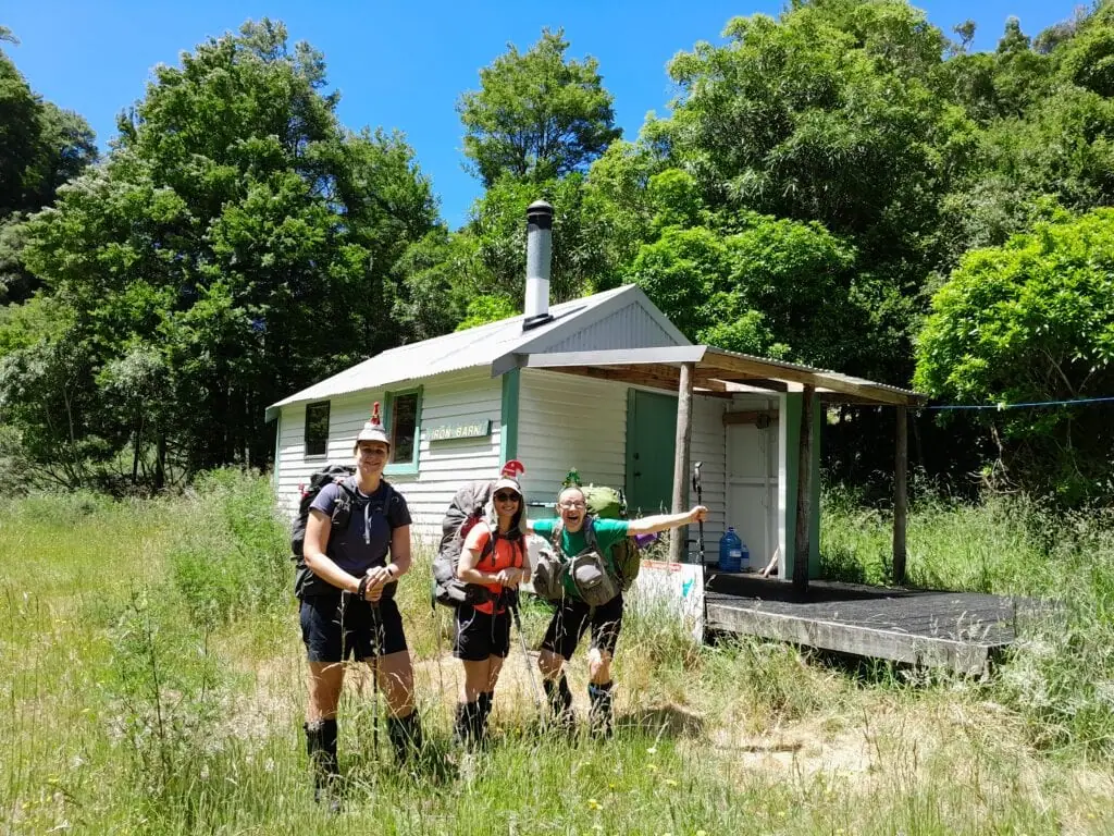 Three smiling trampers wearing Christmas hats standing outside the white timber Iron Bark Hut on a sunny grassy flat.