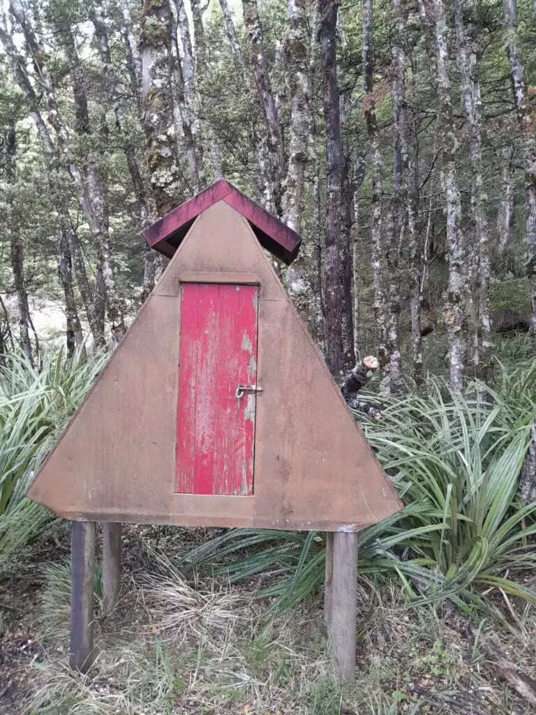 A small a-frame meat safe with a red door at Maropea Forks Hut amongst native bush in the Ruahine Range