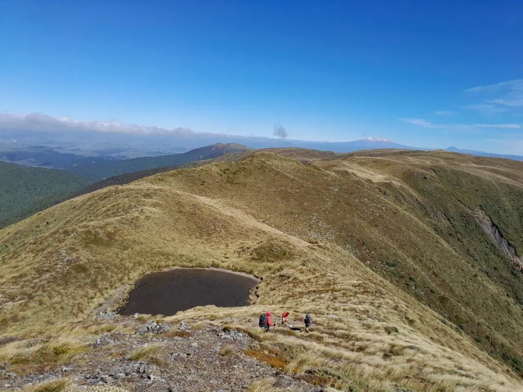 Trampers walking past the tarn below Rongotea peak in a strong wind, surrounded by the deep valleys of the Ruahine mountains with Mt Ruapehu in the distance and the golden tussock of the Mokai Patea Plateau