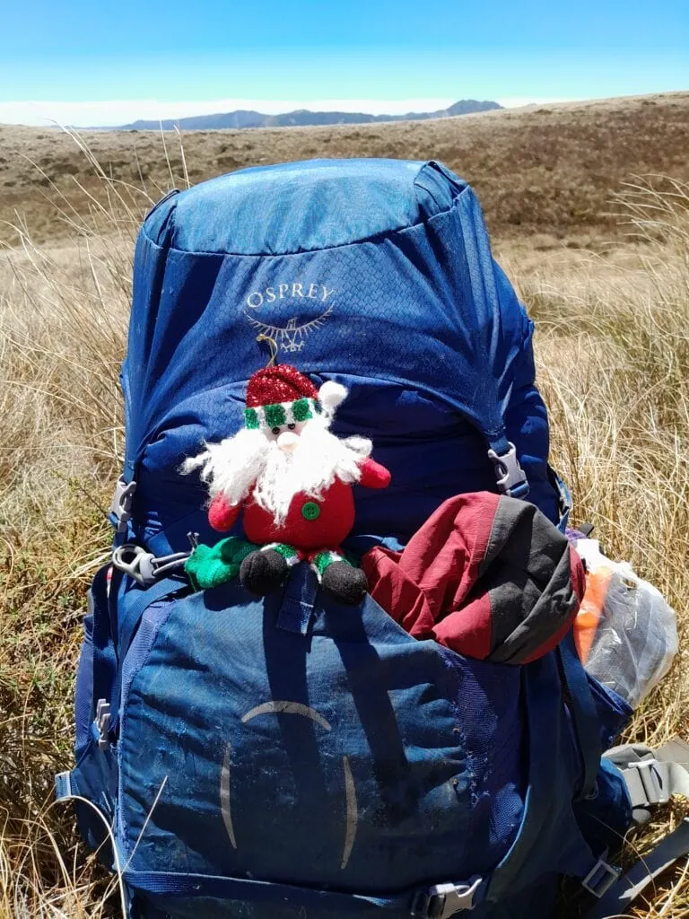 A miniature Santa Claus plush doll hitched onto the back of a tramping pack, with the expansive golden tussock plains of the Mokai Plateau behind.