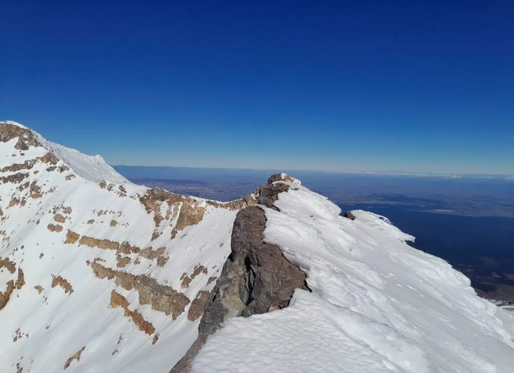 A steep and narrow summit ridge on Mt Ruapehu looking south from Te Ātaahua towards Tahurangi (highest summit) in Tongariro National Park under  a bright blue sky 