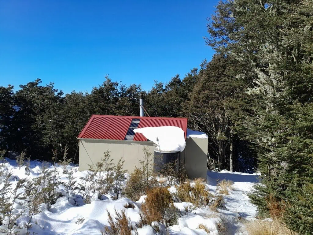 Exterior view of Black Hill Hut with snow on its red corrugated roof, surrounded by native forest, Mt Oxford Forest, Canterbury
