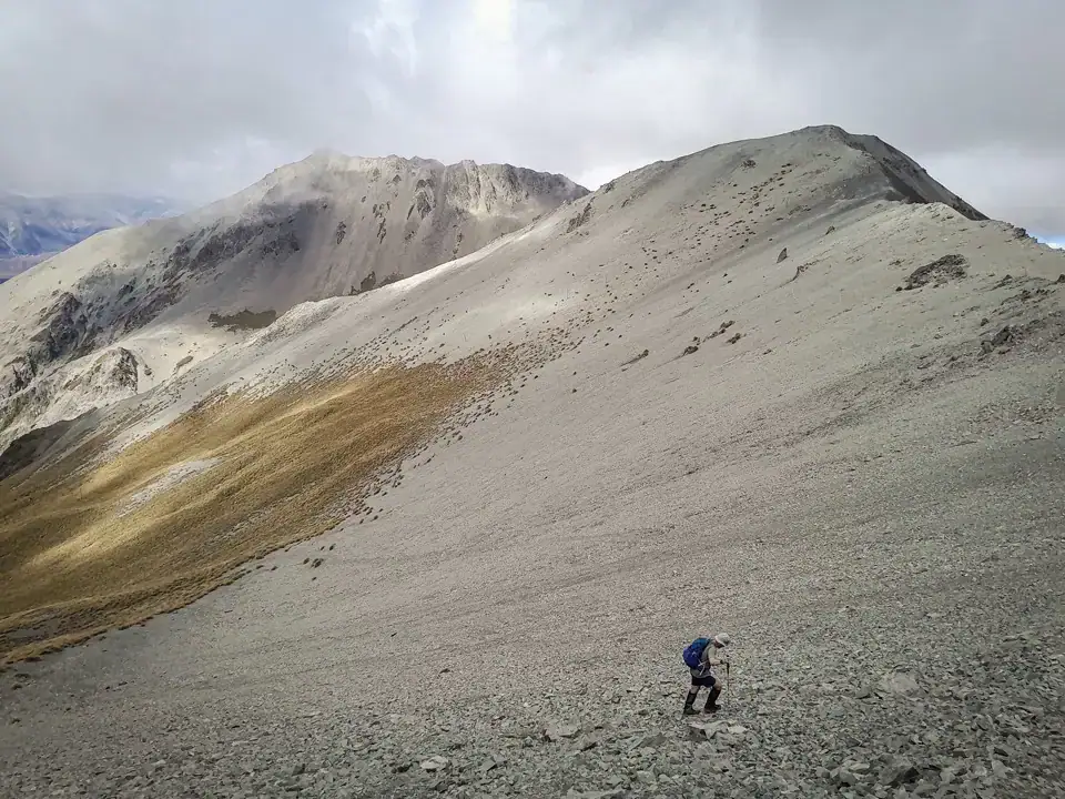 A tramper follows an unformed route along a vast scree slope, passing golden tussock grass in the Broken River Ski Area as they head towards Nervous Knob, a peak of the Craigieburn Range, Craigieburn Forest Park, Canterbury