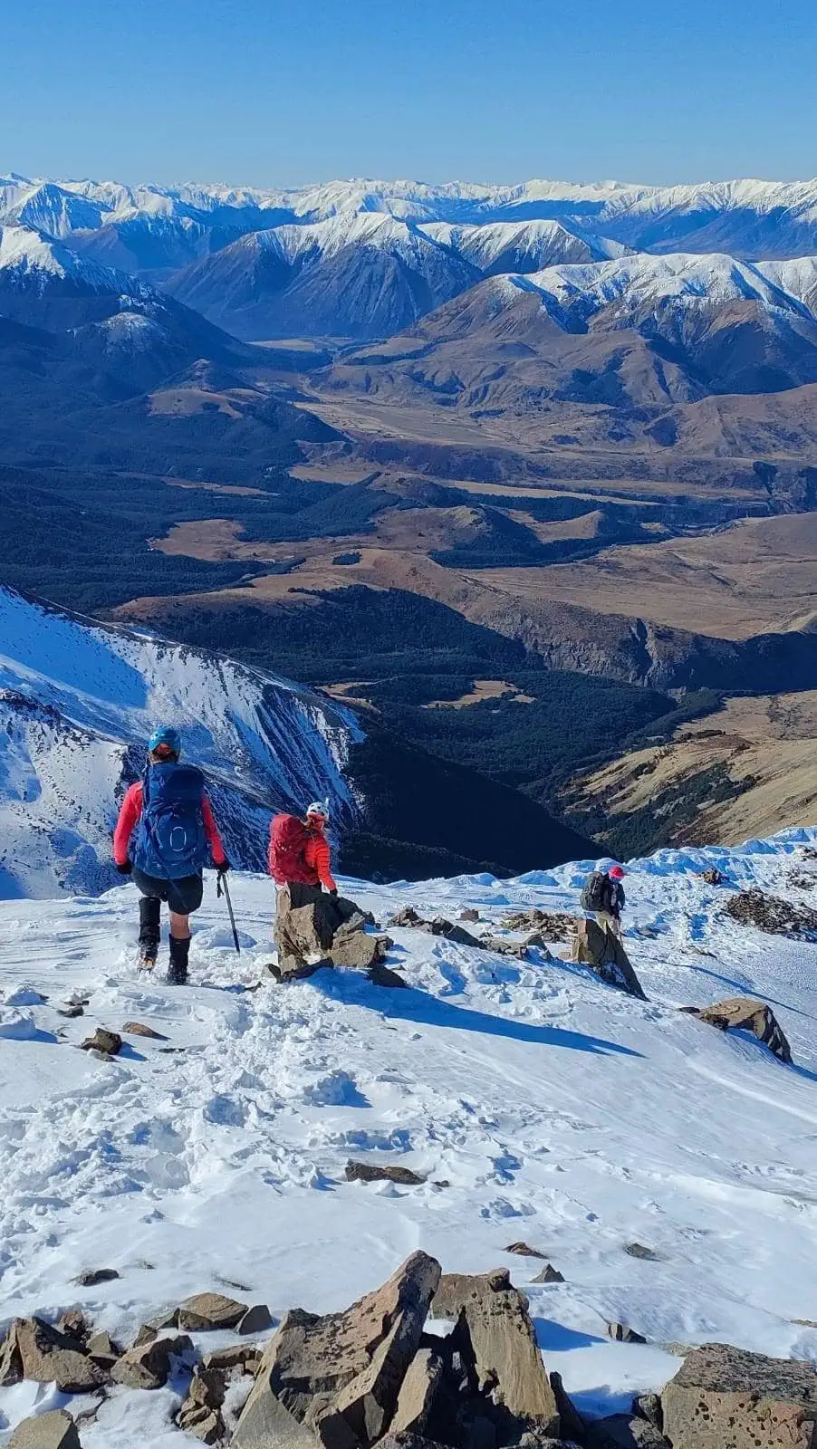 Trampers navigating a steep, snow-covered alpine slope with expansive valley views and across to the Torlesse Range in Craigieburn Forest Park during Canterbury winter.