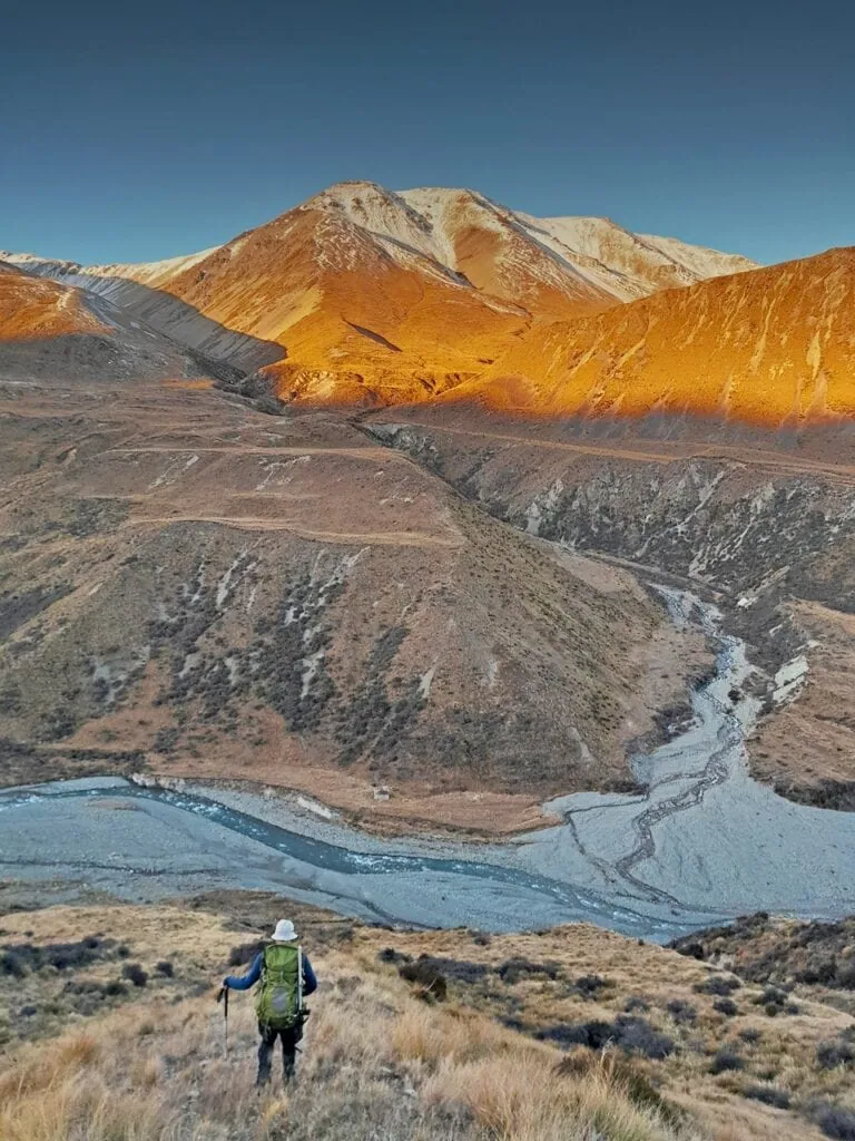 Tramper descending a ridge with a tramping pack during golden hour sunset over Potts River valley and snow-dusted Bill Hill Range in Hakatere Conservation Area