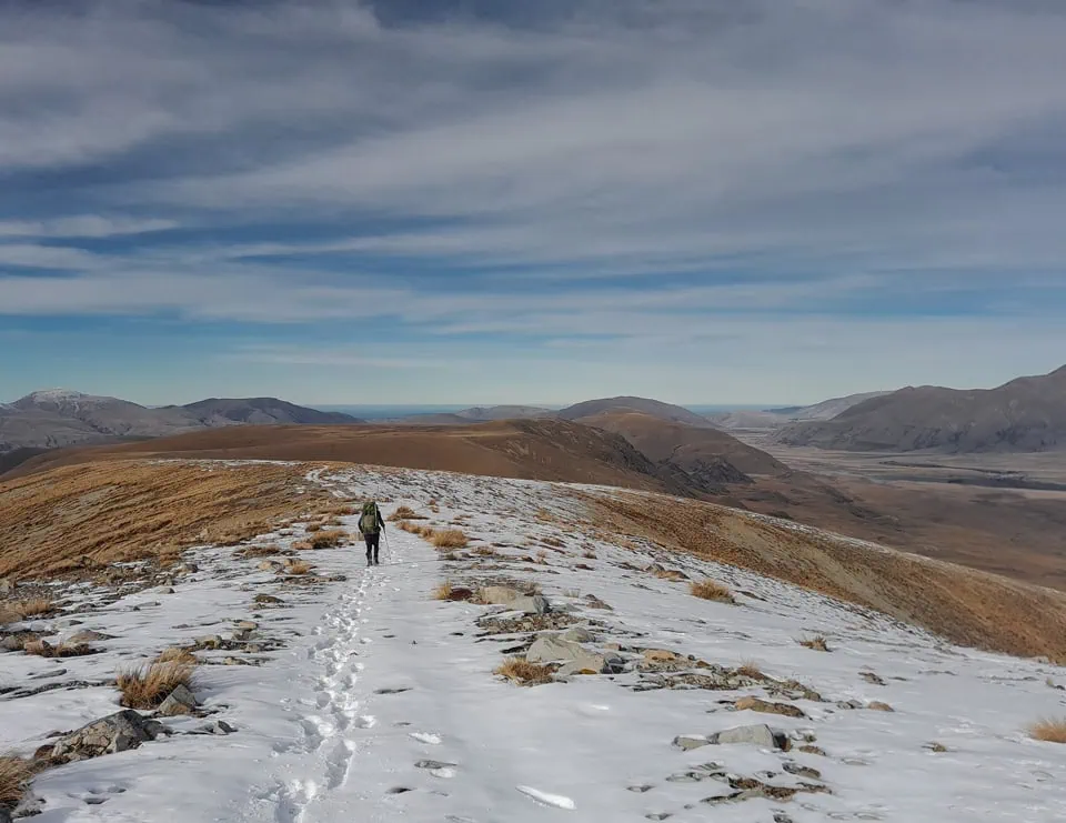 A tramper with a backpack following snowy footprints along a wide alpine ridge on the Mt Potts track on the Dogs Range in winter in Hakatere Conservation Area
