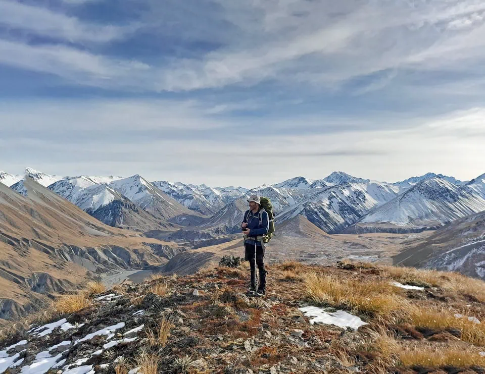 A male tramper with a backpack standing on a rocky alpine viewpoint on the Dogs Range in Hakatere Conservation Area overlooking the vast snow-dusted mountains of Canterbury, New Zealand.