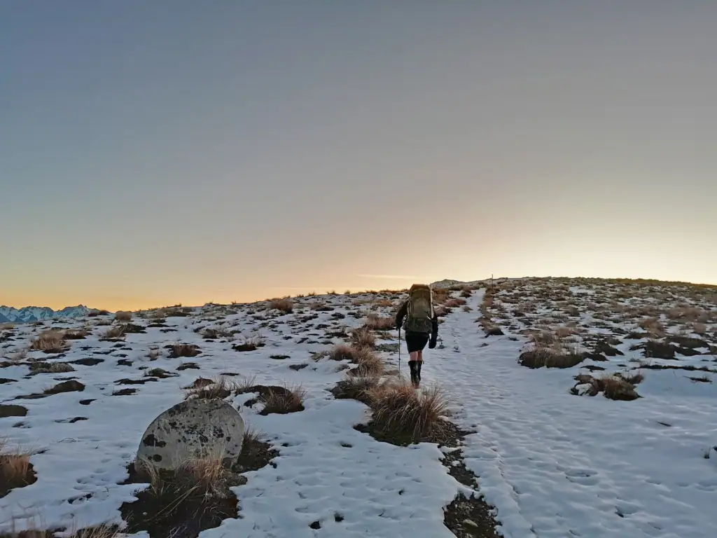 A tramper with a green backpack follows the snow covered Mt Potts track in Hakatere Conservation Park during a pink winter sunset.