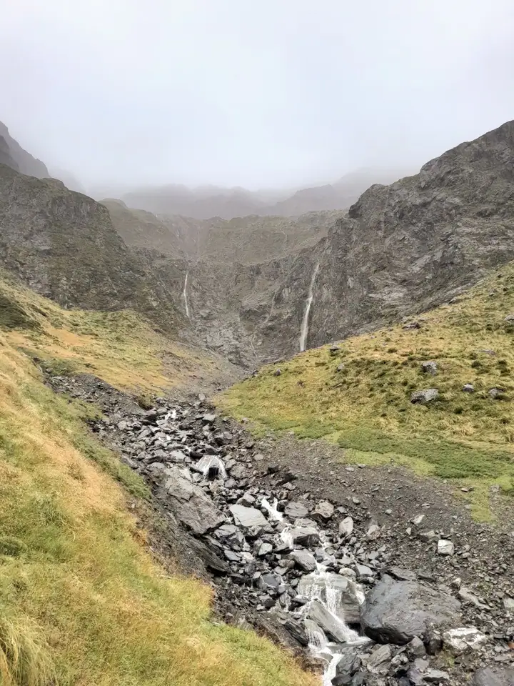 A small mountain stream flowing down a rocky, scree-filled cirque Basin at the head of the North Temple Valley flanked by grassy slopes and waterfalls, Ruataniwha Conservation Park