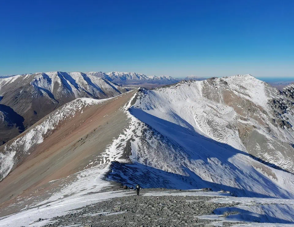 Tramper navigating a snowy alpine ridgeline at just under 2060m on Mt Potts under a clear blue sky in winter, Hakatere Conservation Area, Canterbury High Country