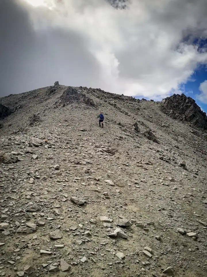 A tramper with trekking poles and a backpack steadily climbs a steep, unformed scree slope towards a rugged, rocky Hamilton Peak, summit in the Craigieburn Range, Craigieburn Forest Park, Canterbury