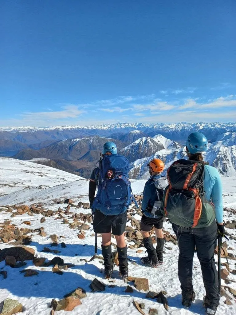 Three trampers wearing climbing helmets and holding ice axes stand on the summit of Mt Izard in Craigieburn Forest Park in winter looking out towards Lake Coleridge