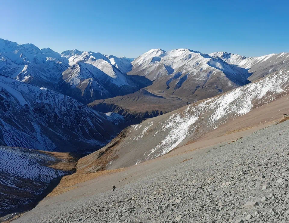 A lone tramper ascending a steep, grey scree slope on Mt Potts with expansive views of snow-covered alpine ranges in Hakatere Conservation Area, New Zealand.