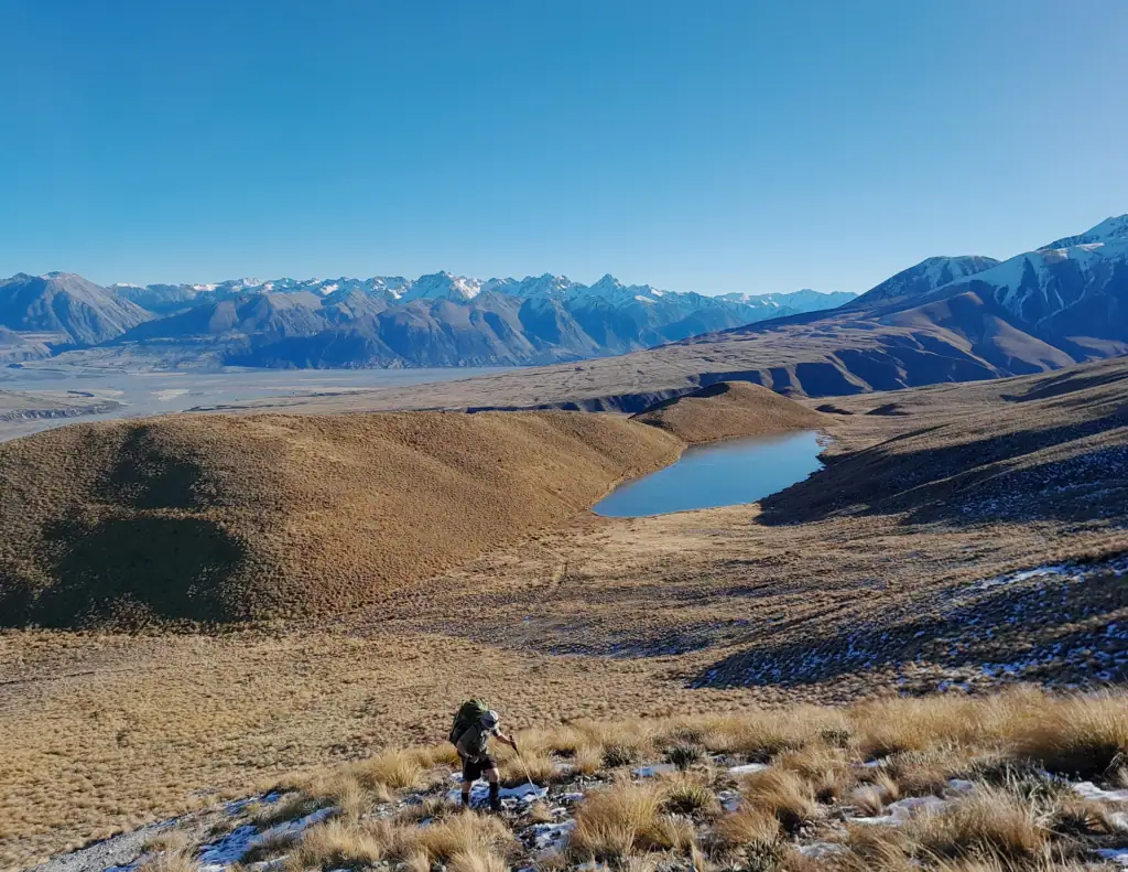 A tramper amongst tussock and snow above Lake Mystery in Hakatere Conservation area, walking into Potts Hut