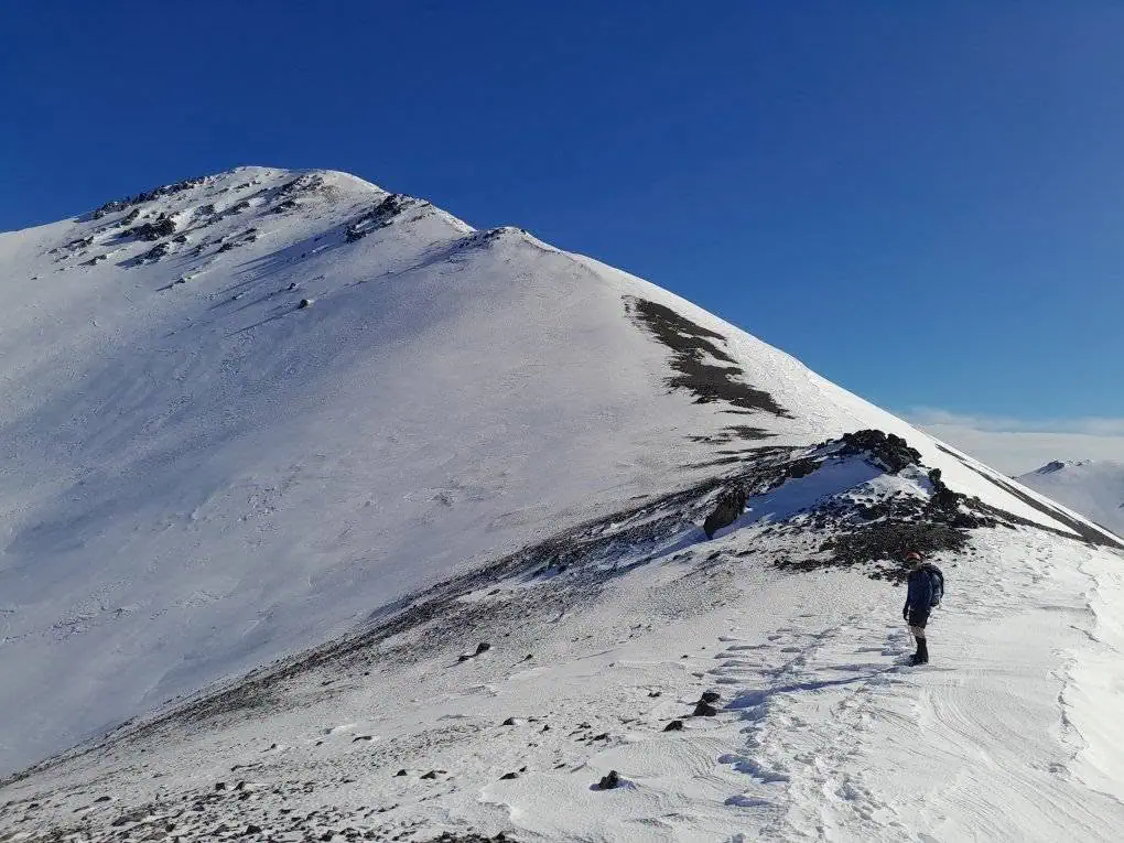 A tramper standing on a snow-covered Long Spur route ascending toward the summit of Mt Cloudsley in the Craigieburn Range under a clear blue sky, Canterbury