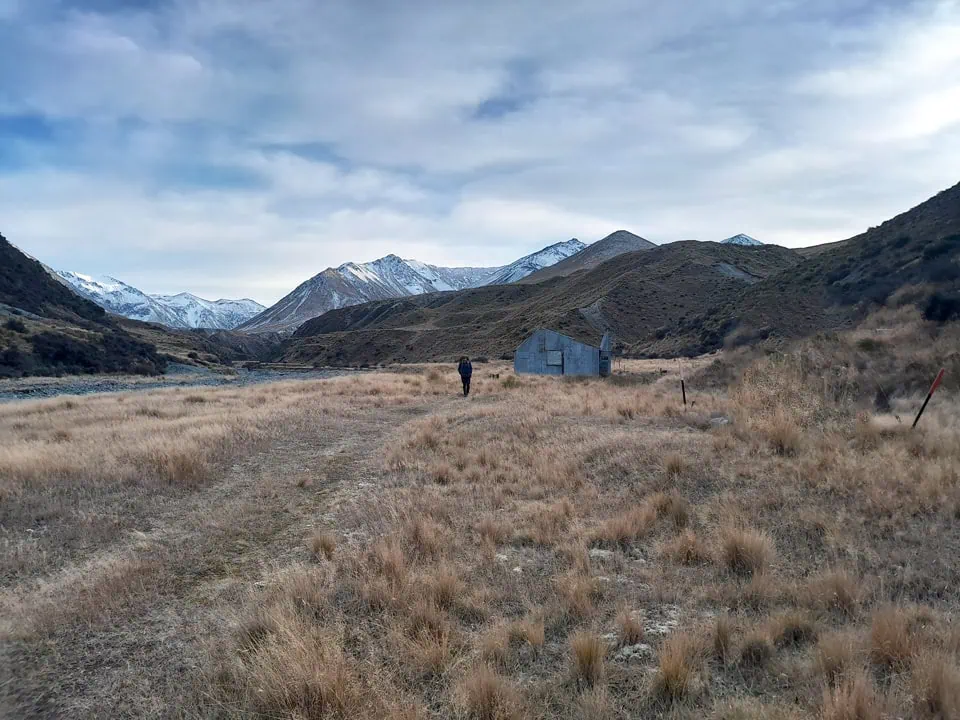 Tramper walking through tussock flats toward Potts Hut in Hakatere Conservation Park with snow-capped Southern Alps in the background