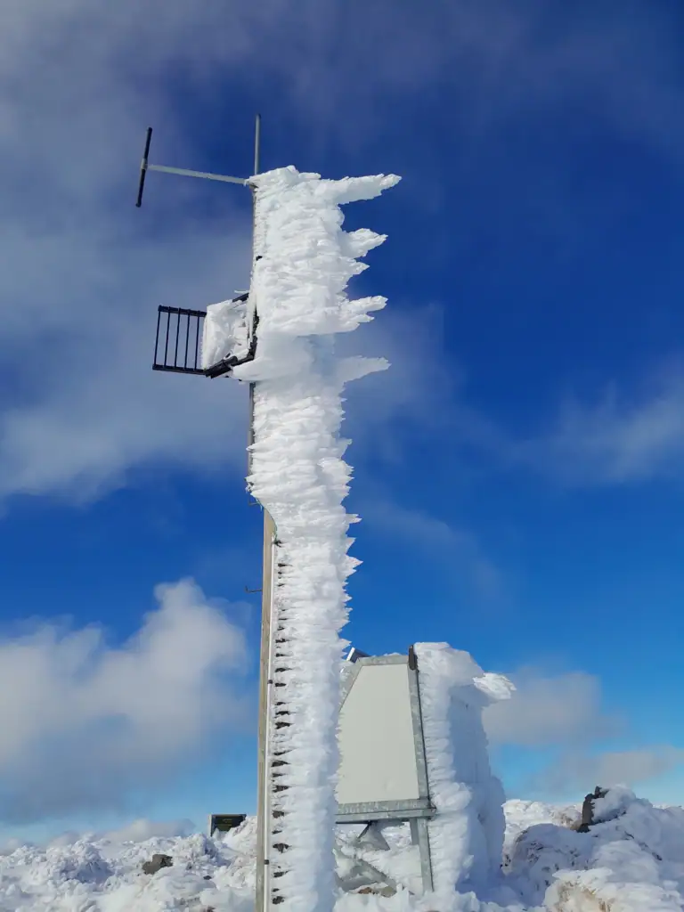 Thick rime ice accumulation coating a communications mast on the frozen summit of Mt Oxford under a blue sky.