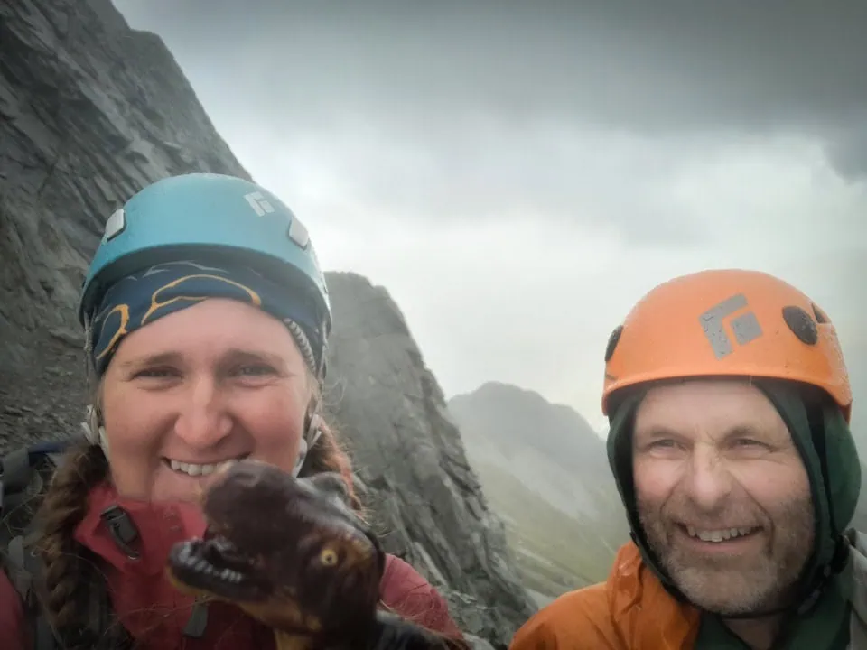 Close-up selfie of two hikers wearing climbing helmets and smiling on the misty mountain saddle of Gunsight Pass with a toy dinosaur in front of them, Ruataniwha Conservation Park
