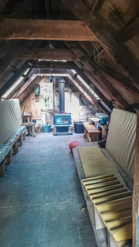 Interior view of the tidy Benmore Hut showing mattress bunks, bench seating, and a wood burner stove under an A-frame timber ceiling