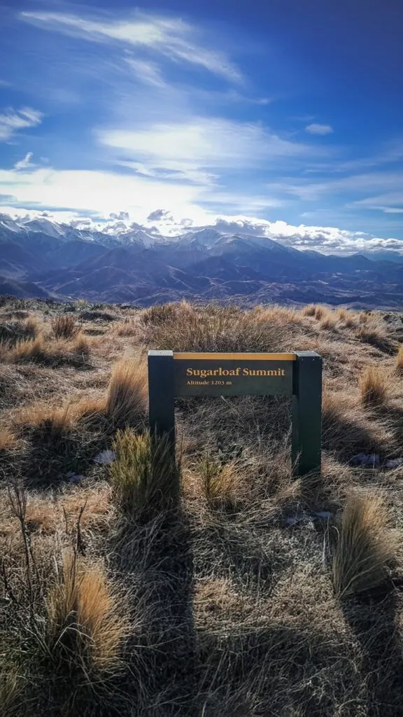 The official green and gold DOC signpost for Sugarloaf Summit, Altitude 1203m, looking out over the Torlesse Range (Canterbury high country) mountain peaks on the Benmore-Annavale Loop