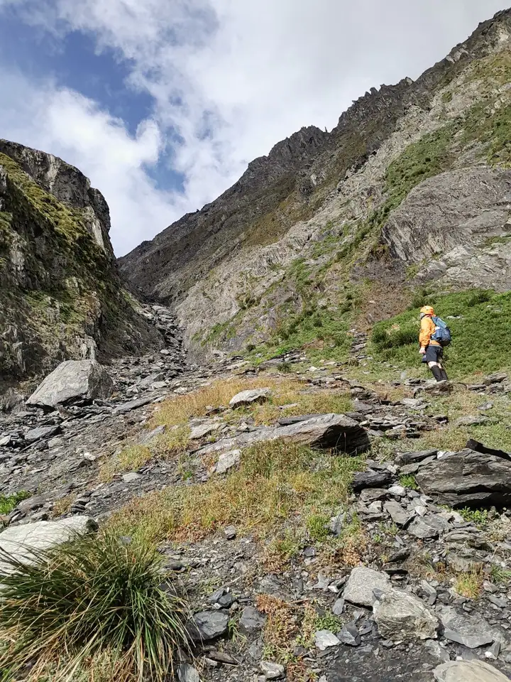 Tramper in an orange jacket and helmet climbing a steep, rocky boulder gully towards Gunsight Pass in Ruataniwha Conservation Park, Canterbury, New Zealand.