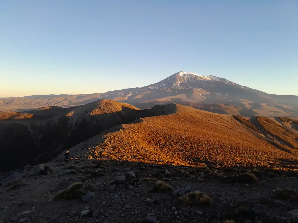 The silhouette of a lone hiker on a steep, dark ridgeline overlooks a vast, arid valley illuminated by a brilliant orange and red sunset, viewed from high above Upper Tama Lake in the Golden Hour in Tongariro National Park