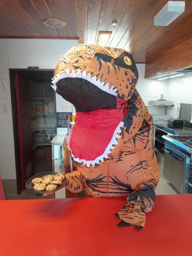 A person wearing an inflatable T-Rex dinosaur costume holding a plate of fresh chocolate chip cookies inside the kitchen of the WTMC Lodge