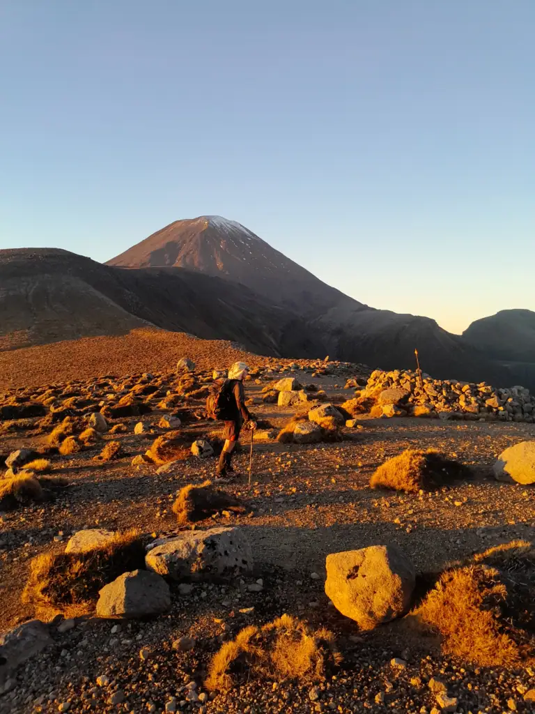 A hiker with a backpack and walking pole explores the rocky Upper Tama Lakes track in Tongariro National Park at sunset, bathed in golden light with a volcanic peak (Mt Ngauruhoe) in the background.