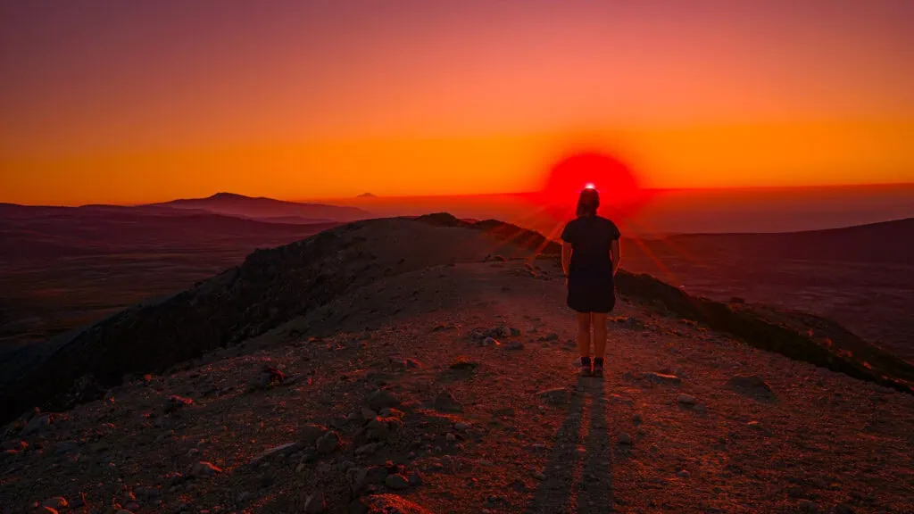 Silhouette of a hiker standing on a high mountain ridge facing a vibrant orange and red sunset flare over the volcanic landscape near Upper Tama Lakes