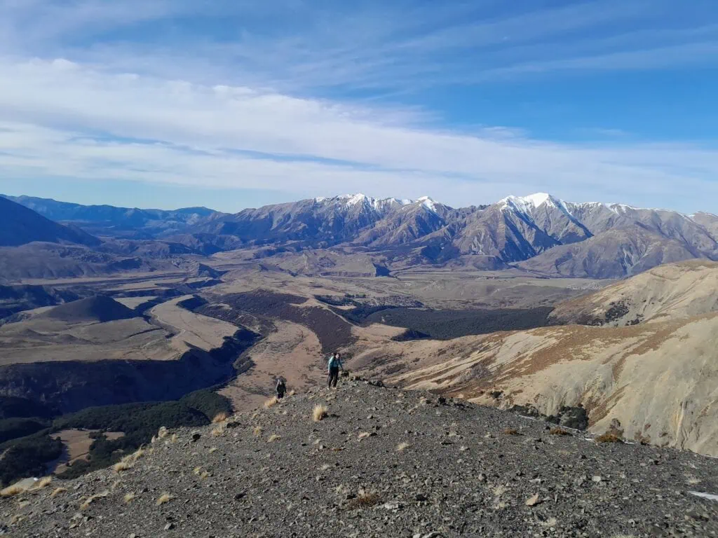 Two hikers ascending a steep rocky scree ridge on the Mt Izard route in Craigieburn Forest Park, looking out over the broad Canterbury high country and distant snow-capped Torlesse Range in the background