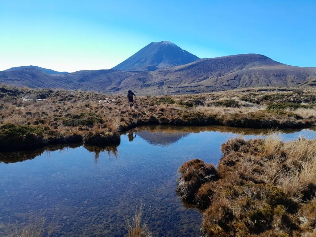 A hiker walks past a still alpine tarn showing a clear water reflection of Mt Ngāuruhoe at Ngā Puna a Tama, the Springs of Tama
