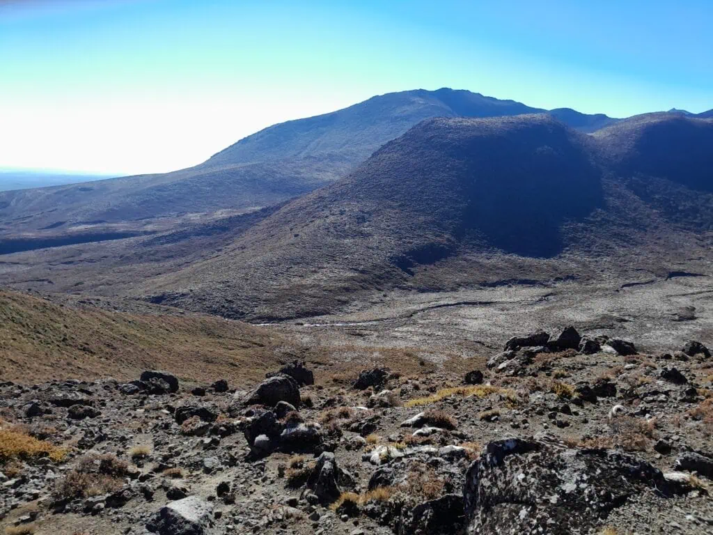 A panoramic view across a dry, rocky alpine valley in the Tongariro volcanic complex, showing the peak of Pukekaikiore in the background
