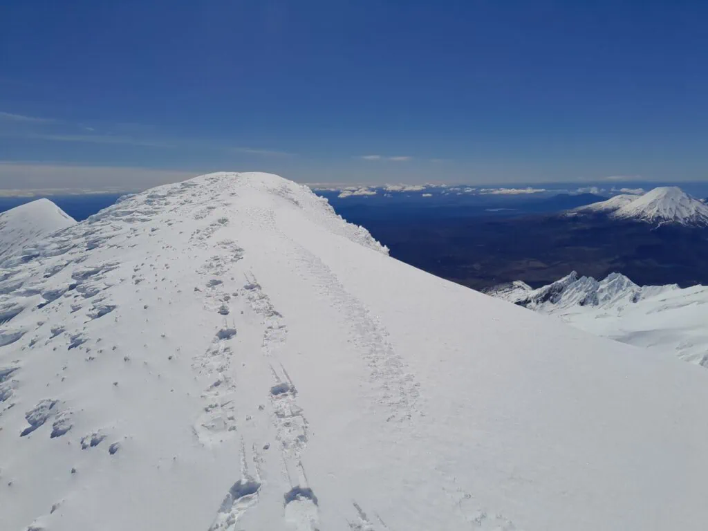 Footprints and crampon tracks leading along the narrow, snow-packed summit ridge of Paretetaitonga with Mount Ngauruhoe visible in the distant background.